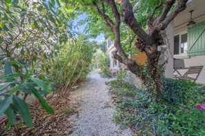 Ein schmaler Kiesweg führt zu einem Gebäude mit einem Balkon, umgeben von üppiger Vegetation und einem Baum.