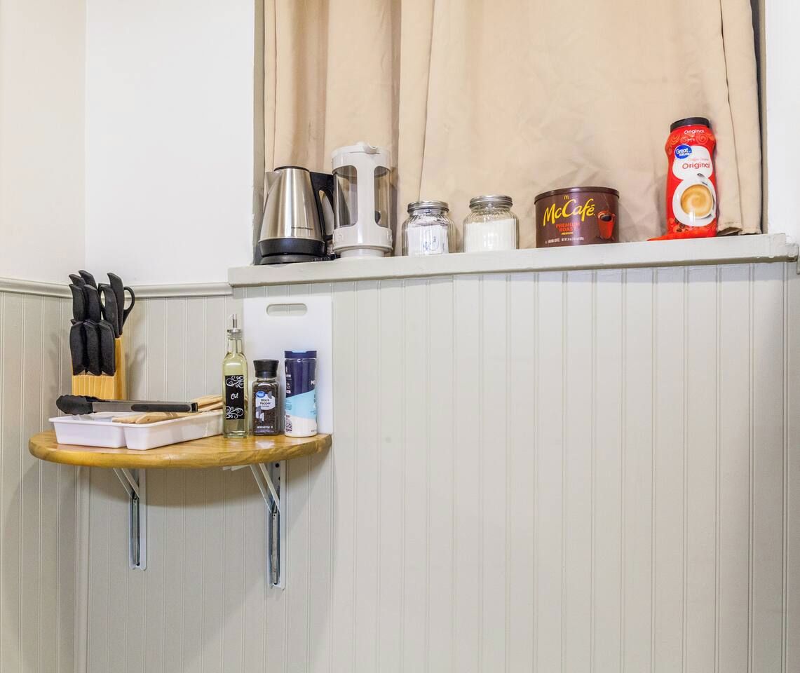 Uma bancada de cozinha com cafeteira, chaleira e uma estante com uma lata de McCafé e um pote de açúcar.