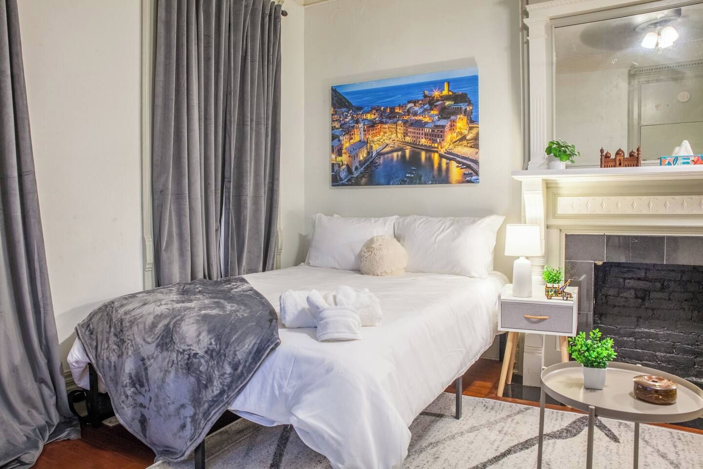 A bedroom with a bed, a painting of a coastal town, a fireplace, and a small table with a plant.
