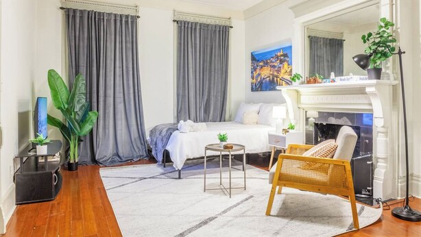 A hotel room with a bed, a small table, a chair, a fireplace, and a painting on the wall. - Modern Studio in Allentown (Buffalo)