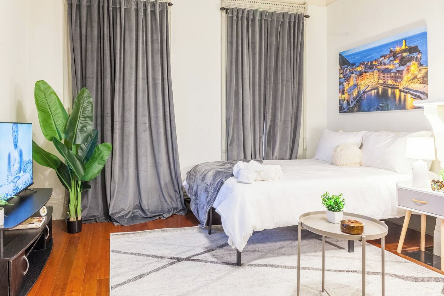 Quarto de hotel com cama, escrivaninha, cadeira, televisão, planta e um quadro na parede.