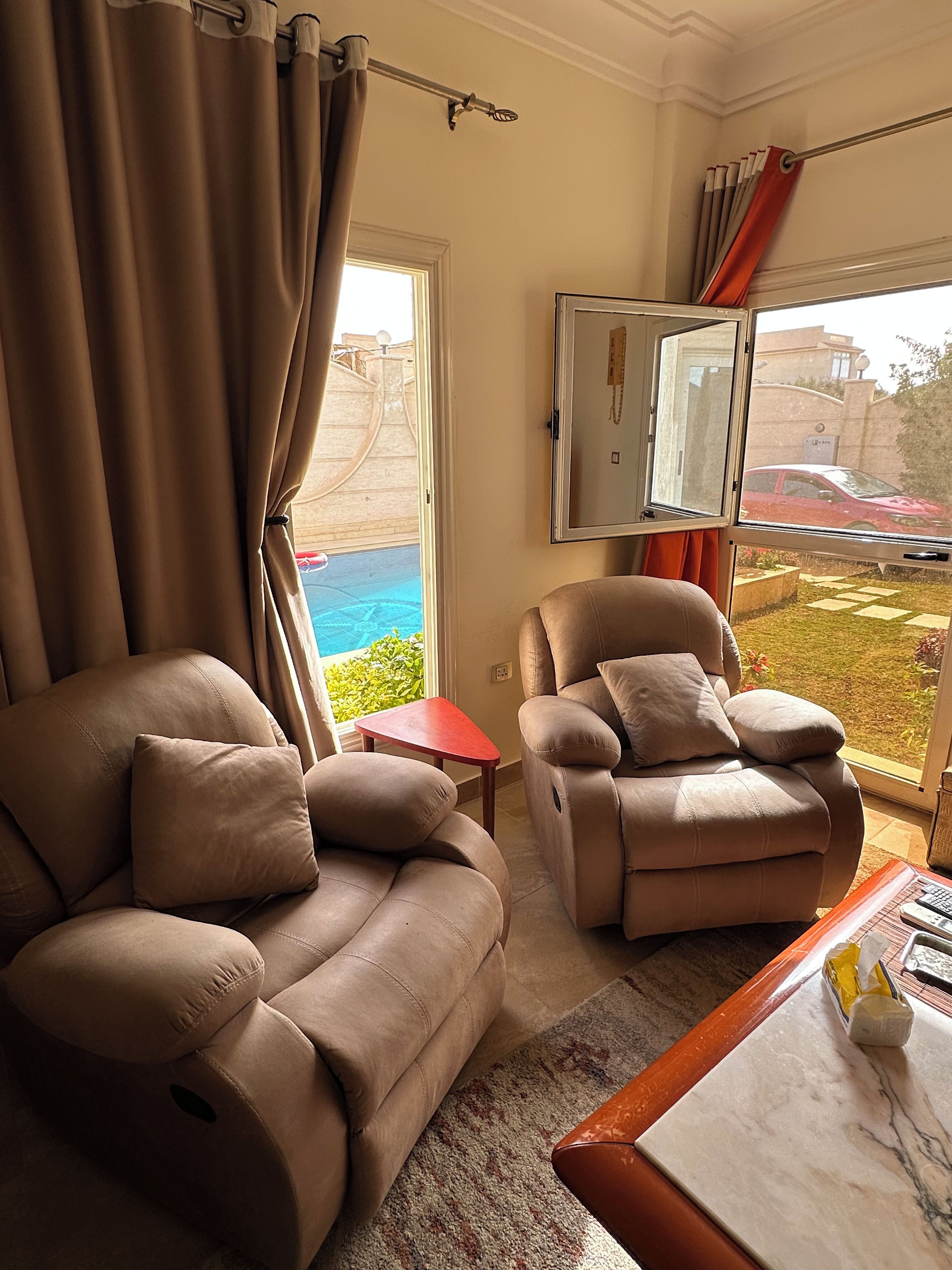 Ein Zimmer mit zwei Liegesofas, einem kleinen Tisch und einem großen Fenster mit Blick auf einen Swimmingpool und ein rotes Auto draußen.