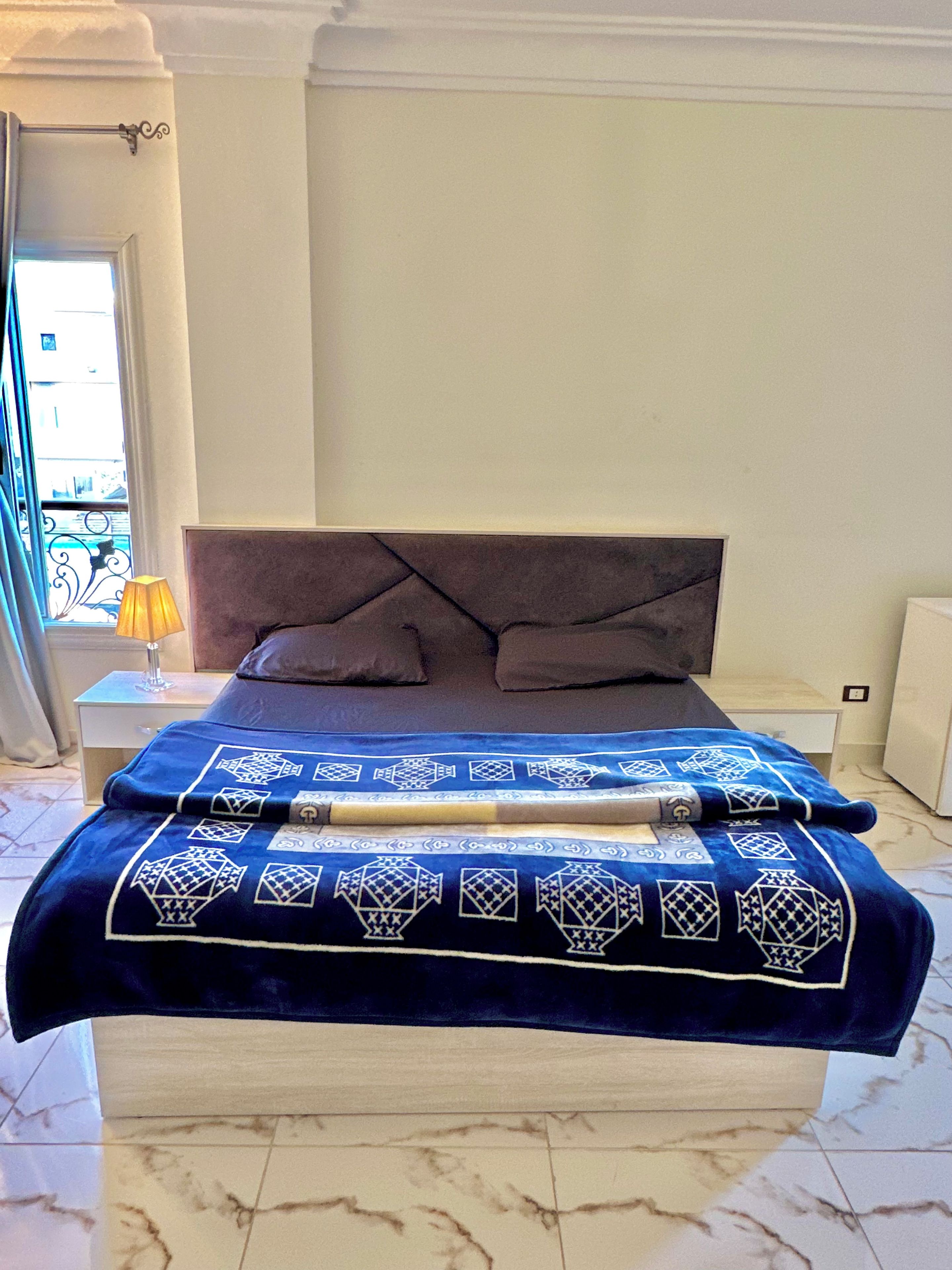 A bedroom with a bed, a nightstand, a lamp, and a view of the outside.