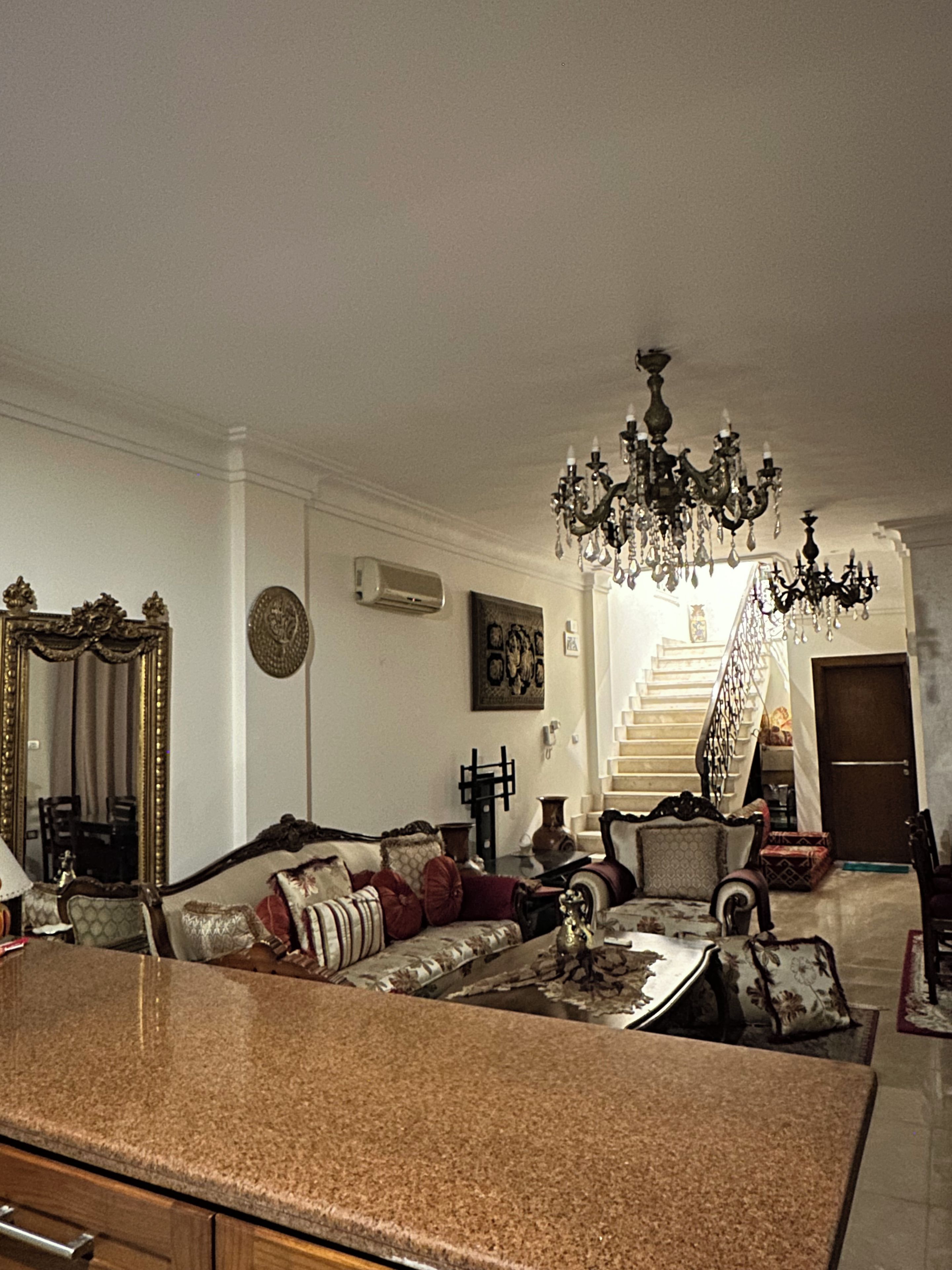 A spacious living room with a grand chandelier, a staircase leading to an upper floor, and a large mirror on the left.