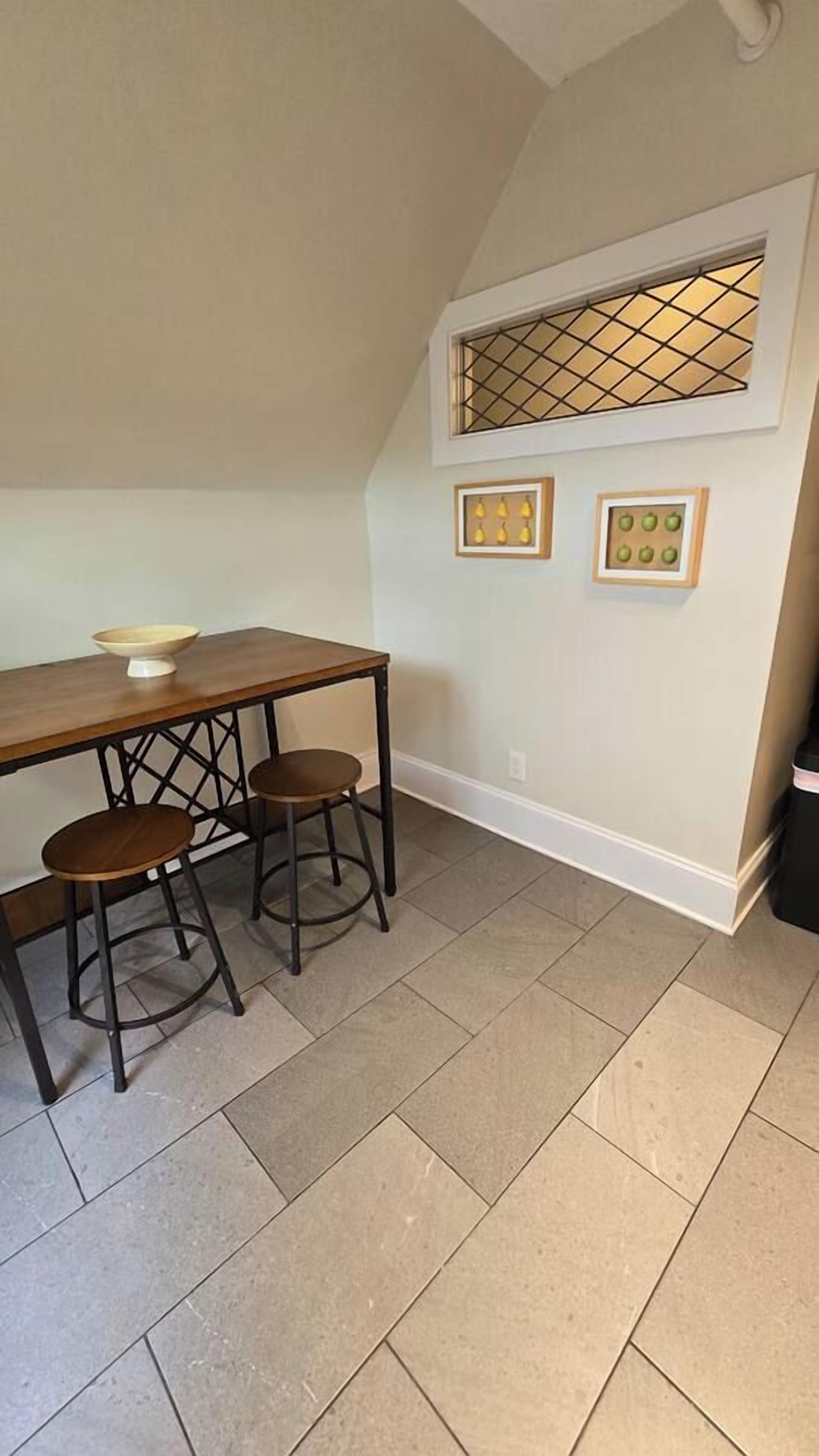 Una cocina con una mesa de madera y dos taburetes, una ventana con diseño de celosía y cuadros en la pared.