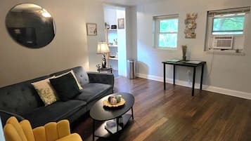Un salon avec un canapé de couleur sombre, un fauteuil jaune, une table basse, un ventilateur de plafond et une fenêtre avec des stores.