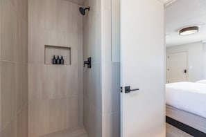 Een hotelbadkamer met een douche, een plank met toiletartikelen en een deur naar een slaapkamer.