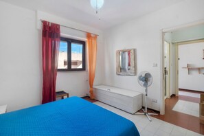 Ein Schlafzimmer mit einem Bett, einem Fenster mit Vorhängen, einem Nachttisch, einem Spiegel, einem Ventilator und einer Tür zu einem anderen Raum.