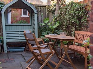 A wooden outdoor seating area with a table and chairs, a small green structure, and a brick wall with plants. - The Royal Exchange House in Canterbury (Kent)