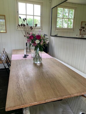 A wooden dining table with a pink placemat, two vases with flowers, and a window with a view of trees. - The Pavilion, Heathfield Lane, West Meon (West Meon)