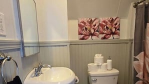 Ein Badezimmer mit einem weißen Waschbecken, einer Toilette und einem Spiegel. Ein schwarzes Handtuch hängt an der Wand, und ein Mülleimer ist sichtbar.