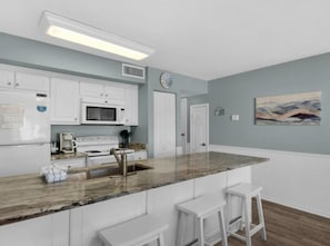 Een keuken met witte kasten, een granieten aanrecht, een magnetron, een fornuis en een spoelbak. Er staan twee witte barstoelen en er hangt een schilderij aan de muur.