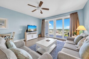 Ein Wohnzimmer mit Deckenventilator, Fernseher, einer Sofagarnitur, einem Couchtisch und Blick ins Freie durch große Fenster.