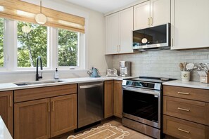 A modern kitchen with wooden cabinets, stainless steel appliances, a built-in microwave, and a window with a view of trees. - The Blue Jetty Coastal Home (Kennebunkport)
