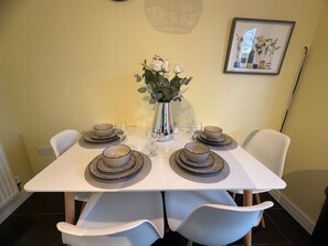 A dining table set with plates, bowls, and glasses, surrounded by white chairs, with a vase of flowers and a framed picture on the wall. - Arnold 3-bed House, Sleeps 5 With Garden & Parking (Nottingham)