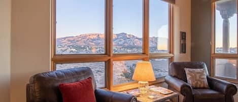 A room with leather sofas, a small table with a lamp, and large windows offering a view of a mountainous landscape.