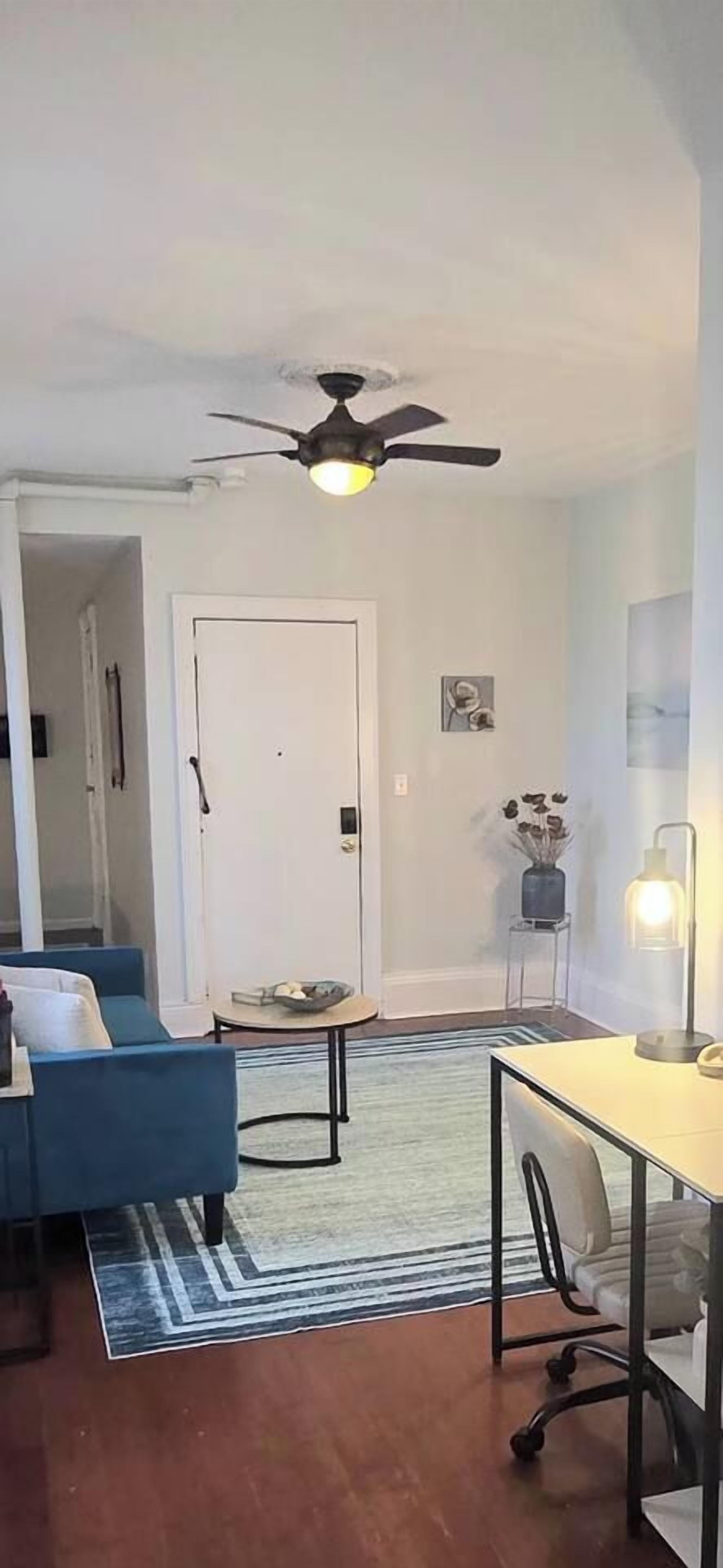 Una sala de estar con un sofá azul, una mesa de comedor, un ventilador de techo y una alfombra.