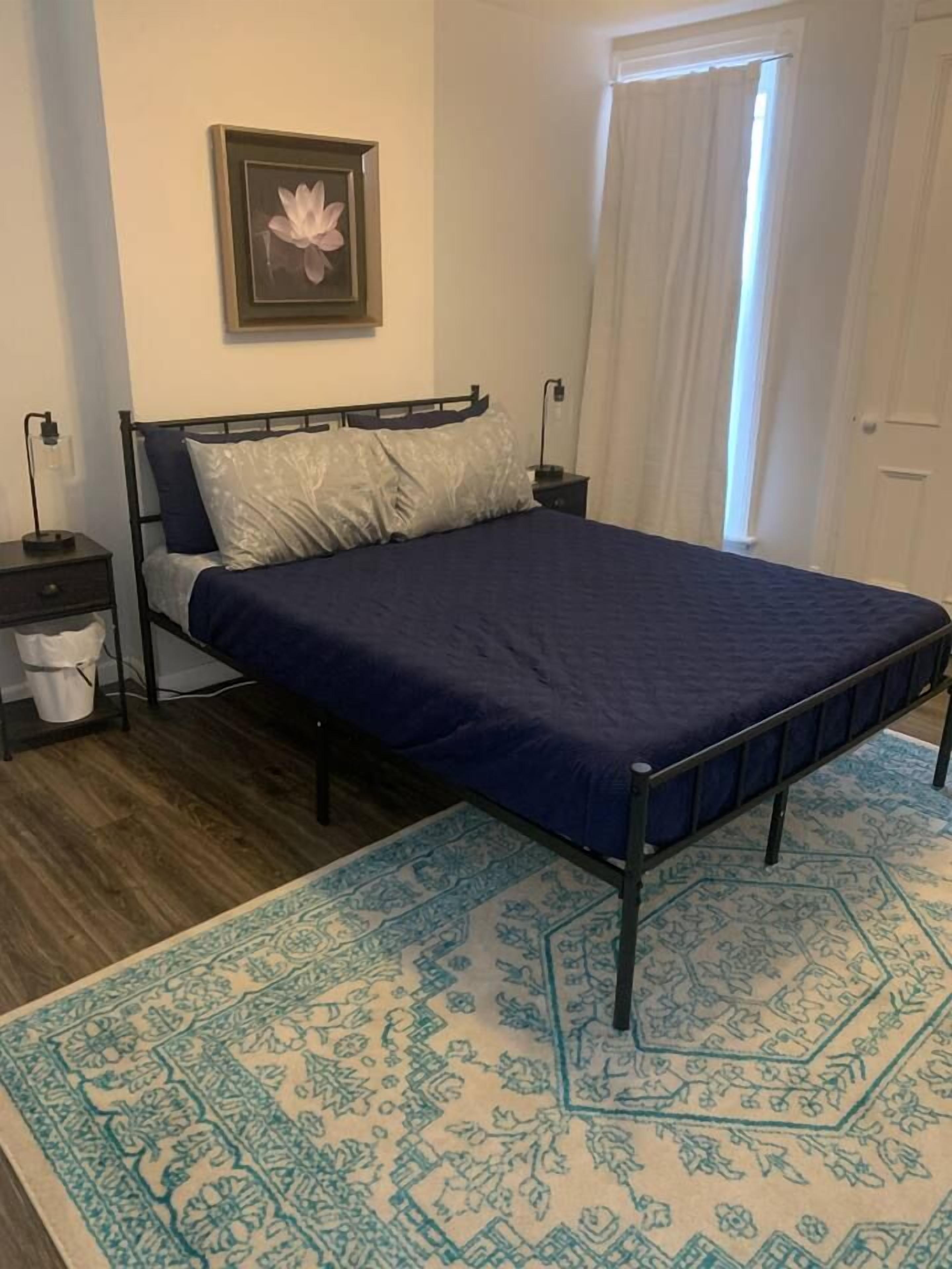 A bedroom with a bed, a nightstand, a wall-mounted picture, and a patterned rug.