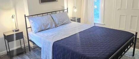 A bedroom with a bed, bedside lamps, a framed picture on the wall, a window with curtains, and a patterned rug.