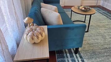 Una sala de estar con un sofá azul, una mesita con una calabaza decorativa y una mesa redonda de centro con un cuenco de fruta.