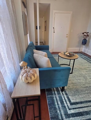 Una sala de estar con un sofá azul, una mesita con una calabaza decorativa y una mesa redonda de centro con un cuenco de fruta.