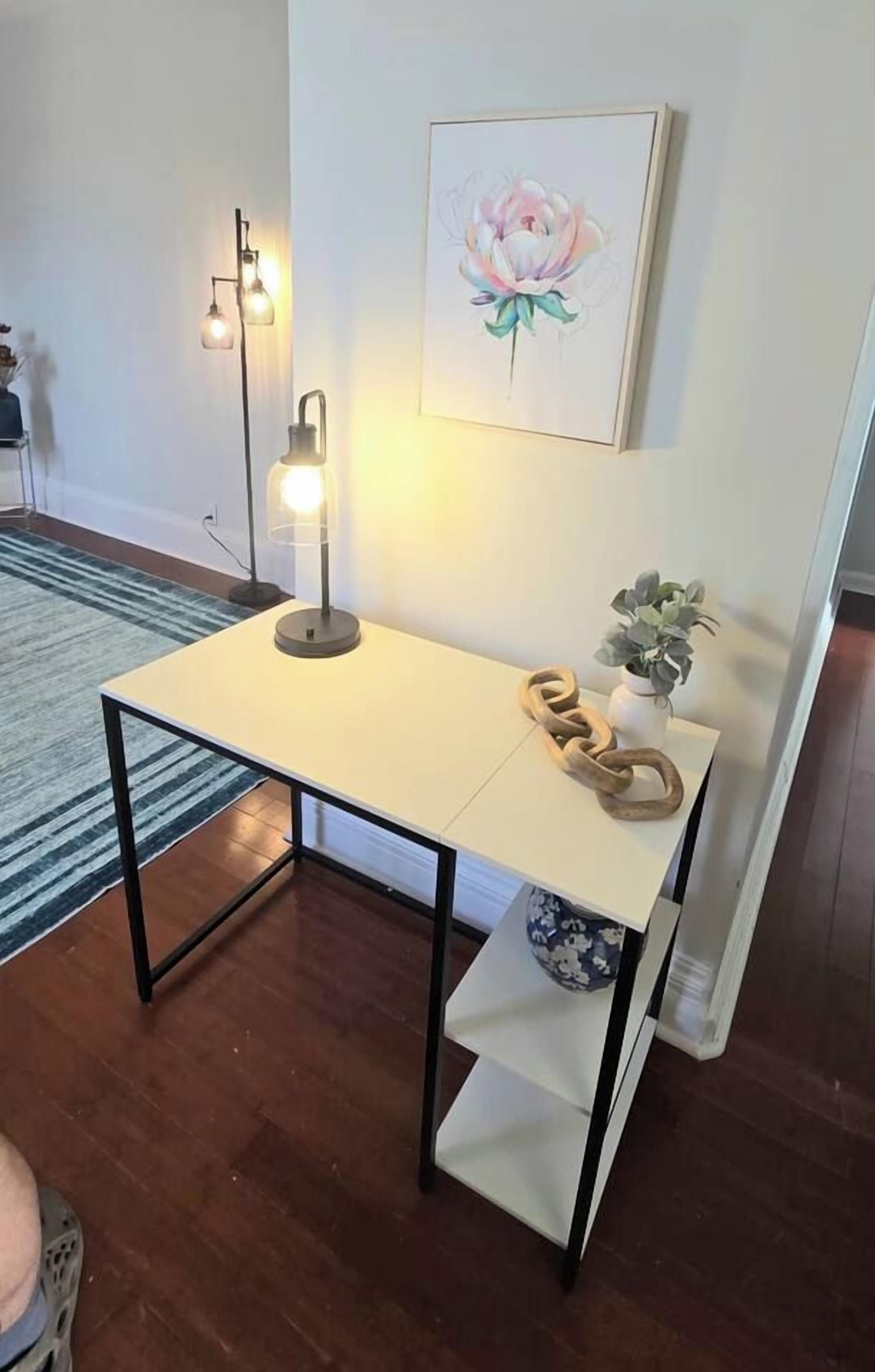 Una habitación con una mesa, una lámpara, una obra de arte enmarcada y una pequeña planta.