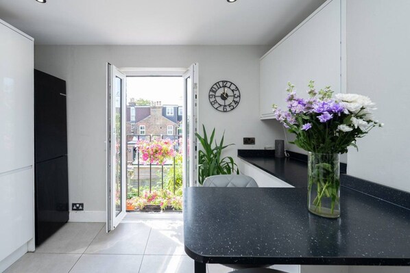 A modern kitchen with a black countertop, a clock on the wall, and a view of a garden through an open door.