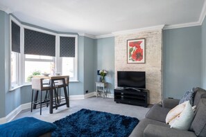 Sala de estar com televisão, sofá, mesa de jantar com cadeiras e tapete azul.