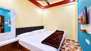 A hotel room with a bed, a desk with a mirror, a TV mounted on the wall, and a patterned floor. - Shree Bada Ganesh Yatri Grah (Ujjain)