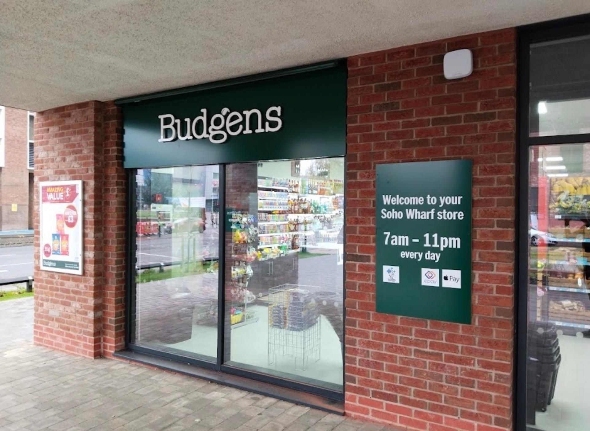 Een winkelgevel met een groen luifel waarop 'Budgens' staat en een bord met de openingstijden.