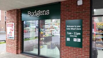Een winkelgevel met een groen luifel waarop 'Budgens' staat en een bord met de openingstijden.