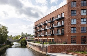 A modern multi-story building with balconies, a river in the foreground, and a bridge in the background. - Luxury 1 Bedroom Apartment Sleeps 2 With Balcony (Birmingham)