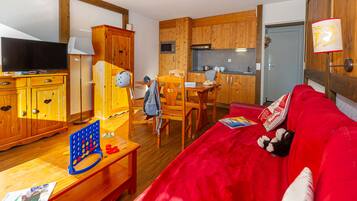 A cozy living room with a red sofa, wooden furniture, a television, and a kitchen area with wooden cabinets and a microwave.