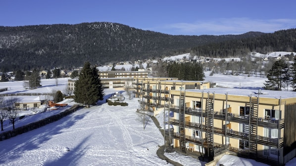 A multi-story building with balconies, surrounded by snow-covered ground and trees, with a mountainous backdrop.