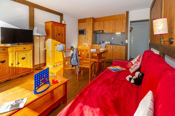 A cozy living room with a red sofa, wooden furniture, a television, and a kitchen area with wooden cabinets and a microwave.