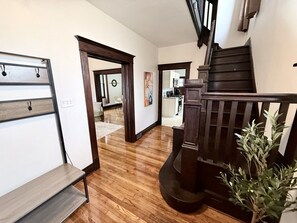 Un escalier en bois menant à l’étage supérieur, un couloir avec un banc, et une vue sur une partie de la cuisine.