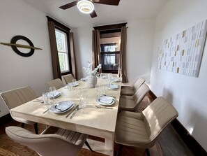 Ein Esszimmer mit einem langen Tisch, der für sechs Personen gedeckt ist, beige Stühle und einer Vase mit weißen Blumen.