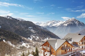 Eine Berglodge mit mehreren Holzhäusern, einer Skipiste im Hintergrund und schneebedeckten Gipfeln.