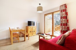 A living room with a red sofa, a wooden dining table with chairs, a TV on a cabinet, and a large window with a view of a snowy landscape. - Au cœur de Vaujany | Piscine | Télécabine à 600 m (Vaujany)