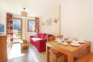 A cozy living room with a red sofa, wooden dining table set with white dishes, and a view of a mountainous landscape through the windows. - À 600 m de la télécabine | Piscine intérieure (Vaujany)