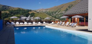 En swimmingpool med liggestole og parasoller, omgivet af et bjergrigt landskab.