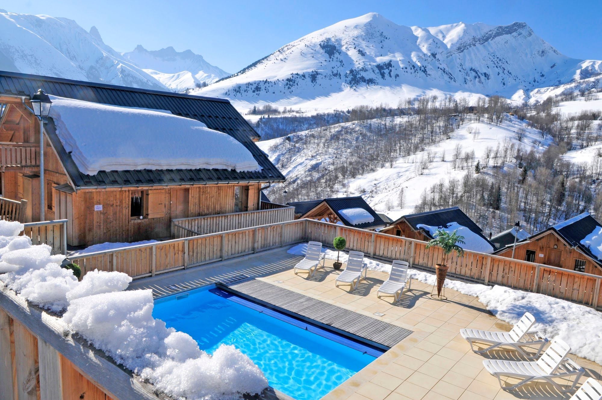 A wooden chalet with a pool and lounge chairs, surrounded by snow-covered mountains.