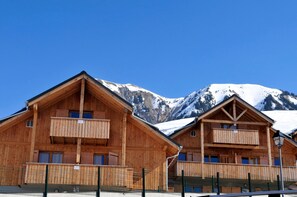 Deux cabanes en bois dotées de balcons, se dressant sur un fond de montagnes enneigées, sous un ciel bleu dégagé.