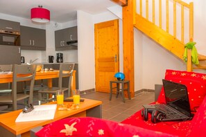 Un salón acogedor con sofá, una mesa con una laptop y audífonos, un comedor con sillas y una mesa, y una cocina con armarios y una cafetera.