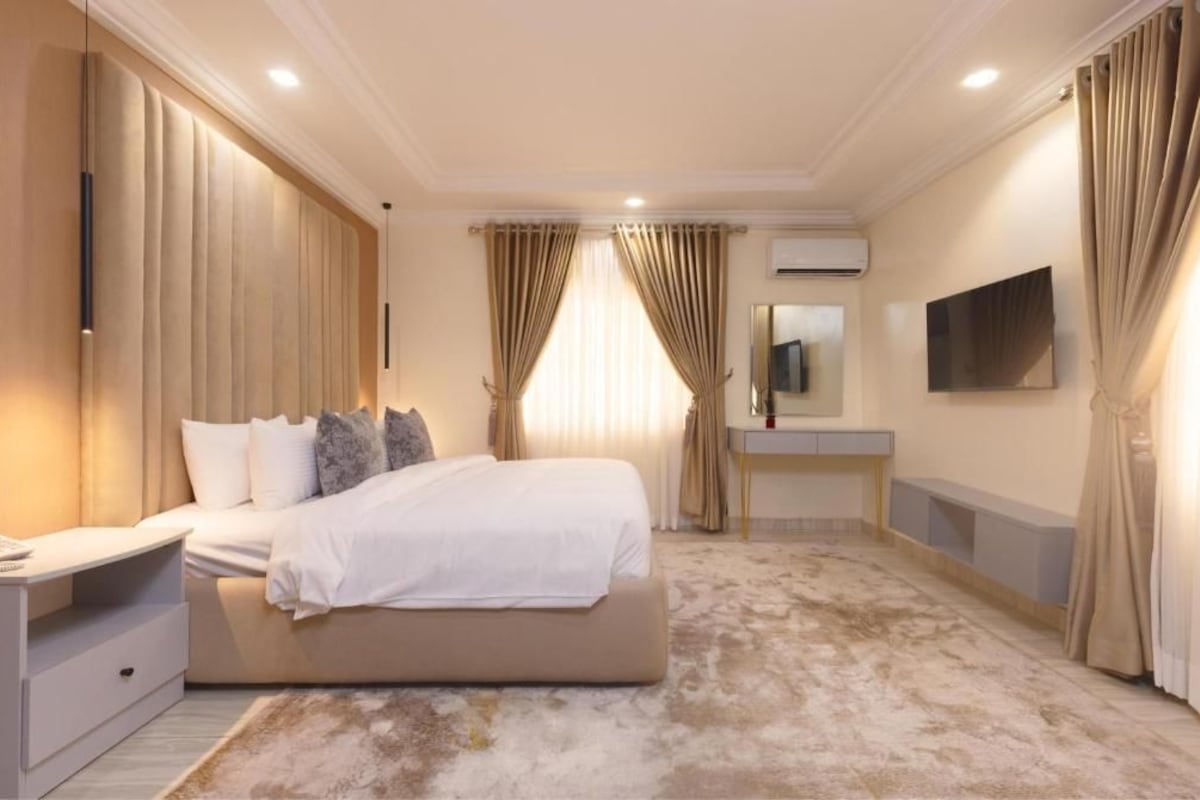 A bedroom with a bed, bedside table, curtains, an air conditioning unit, a mounted TV, and a bathroom vanity.