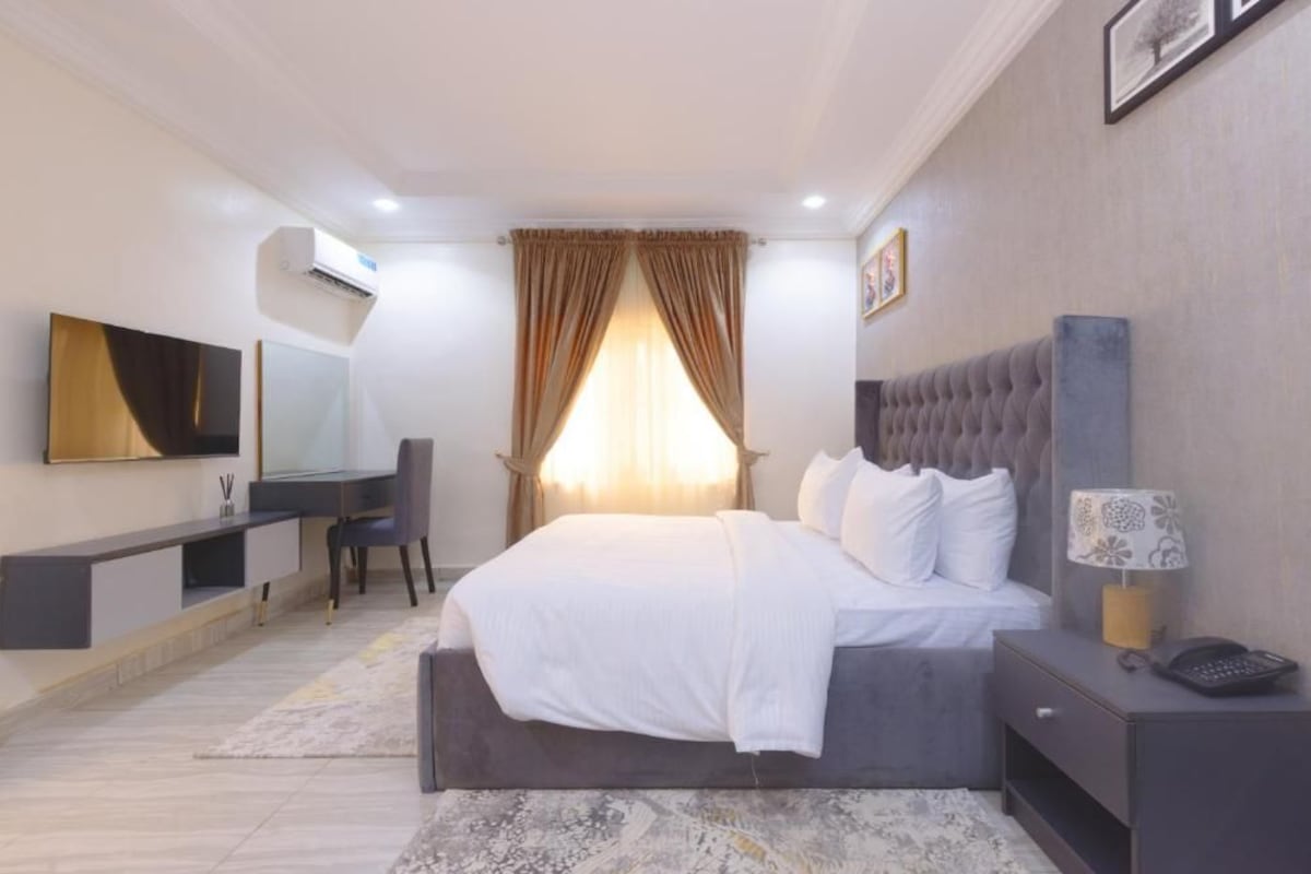 A modern bedroom with a large bed, a desk, a chair, a TV, and a bedside table with a lamp.