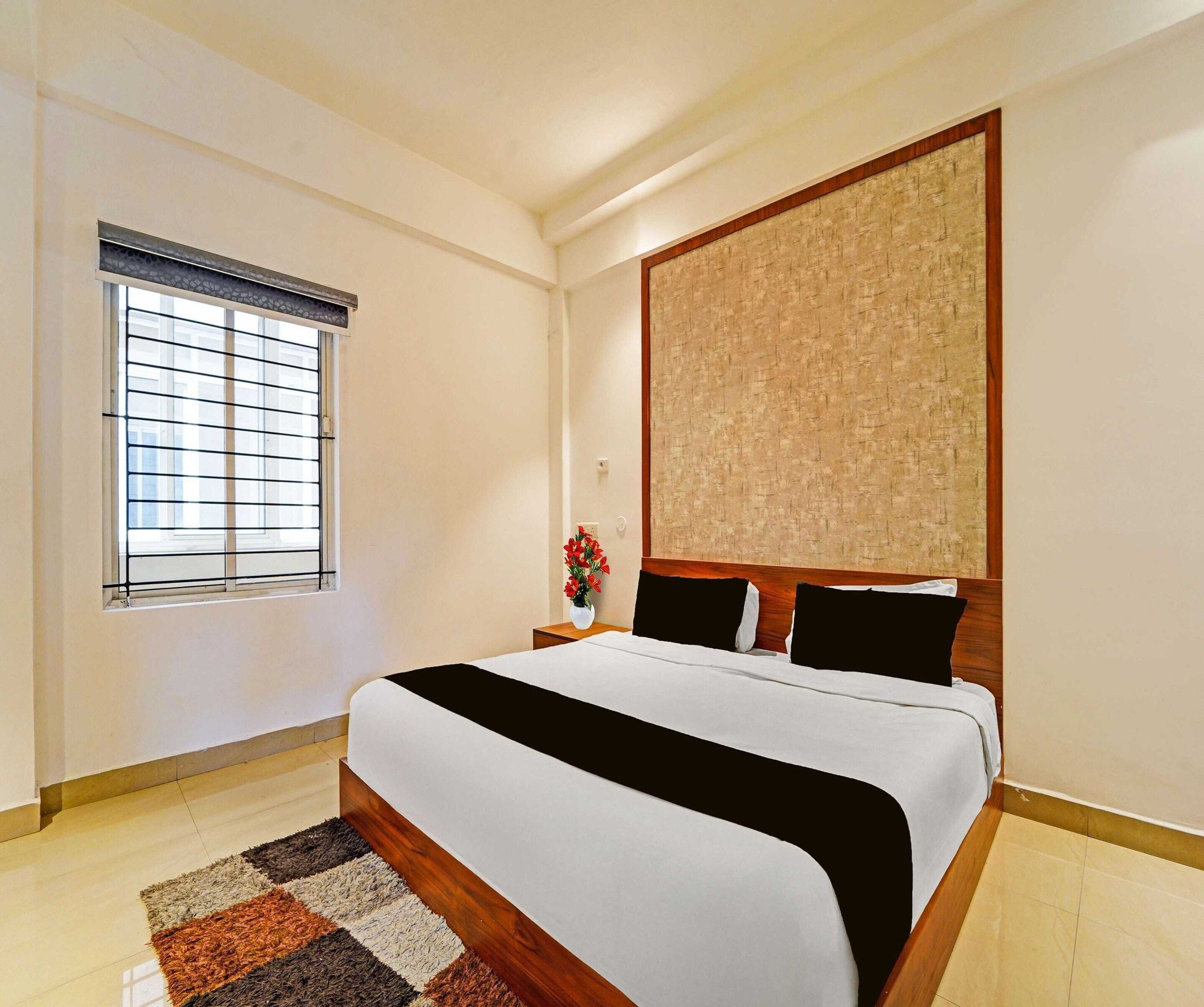 A hotel room with a large bed, a window with blinds, a nightstand with a vase of flowers, and a textured wall panel.