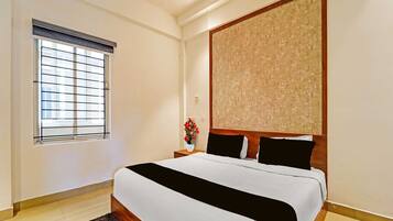 A hotel room with a large bed, a window with blinds, a nightstand with a vase of flowers, and a textured wall panel.