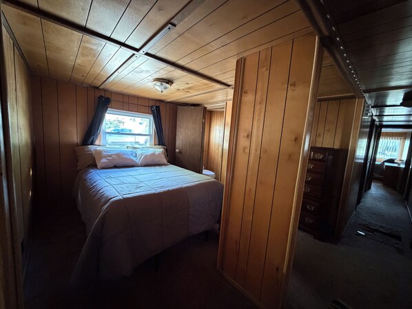 A room with a bed, wooden walls, a window with curtains, and a door leading to another room.