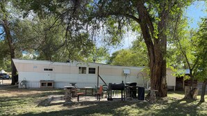 Una caravana amb mobles d'exterior, una barbacoa i un arbre al primer pla.